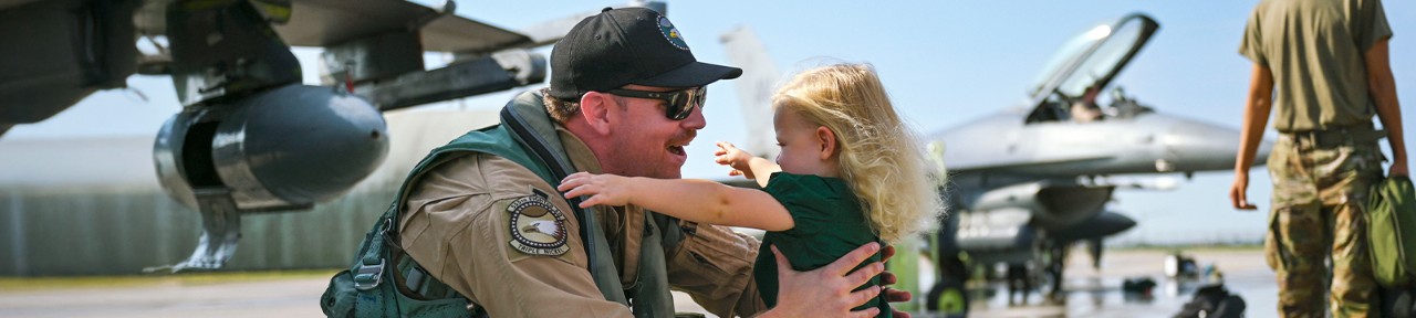 Smiling service member in uniform embraces a child during homecoming. 